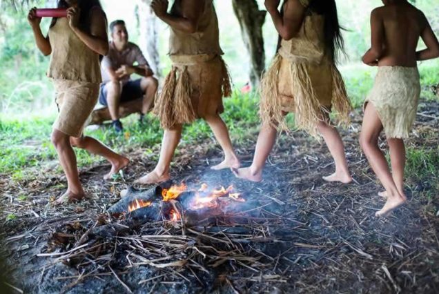 Group of individuals in Tocu Marama skirts dancing around a small fire outdoors.