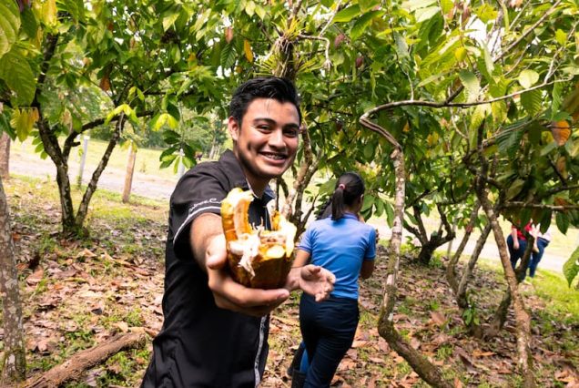 In a lush green orchard in Costa Rica, a person smiles while holding an open cacao pod as part of the Didactic Costa Rica Farm School Tour. In the background, another participant explores the trees with curiosity.