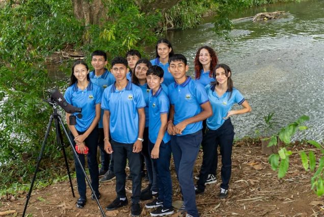 A group of nine young people in blue shirts are gathered by a riverbank during the Didactic Costa Rica Farm School Tour, with a telescope on a tripod beside them. The scene is framed by trees and water, perfectly encapsulating the natural beauty of Costa Rica.