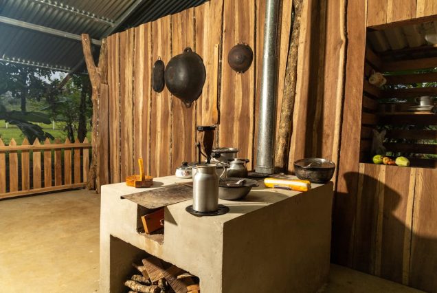 A rustic kitchen with a concrete stove, wood panels, hanging pans, cookware, and a chimney. It has logs stored underneath and a window with shelves on the right.