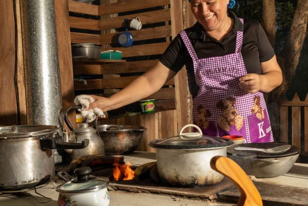 A person in a blue bandana and purple apron cooks with multiple pots and pans on a hot stove in a rustic kitchen.