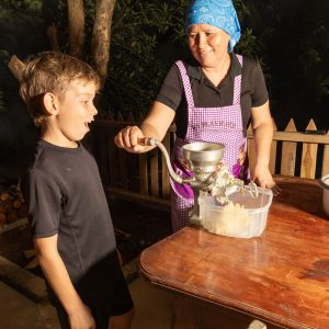 A woman in an apron uses a hand-crank grinder to make food while a young boy watches with interest. They are outdoors near a wooden fence.