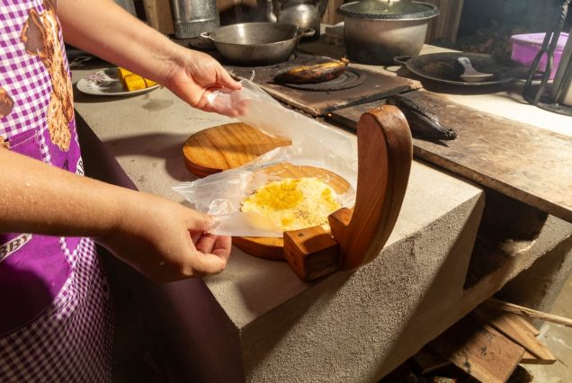 Person presses a corn dough ball using a wooden tortilla press in a rustic kitchen setting.