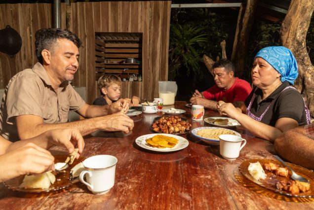 A group of people eating a meal at a wooden table with various dishes, including plantains and sausages, in an outdoor setting.