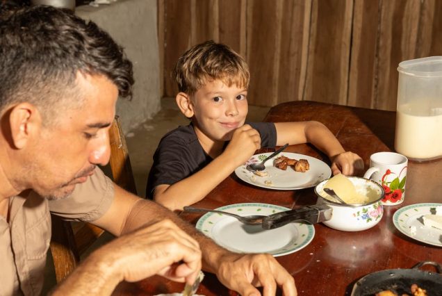 A man and a boy sit at a wooden table eating a meal. The table is set with plates, a cup, and a jug. The boy smiles at the camera.