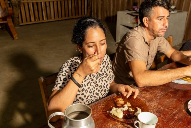 A woman in a leopard-print shirt eats at a wooden table with a plate of food. A man sits beside her. A metal jug and a cup are on the table.