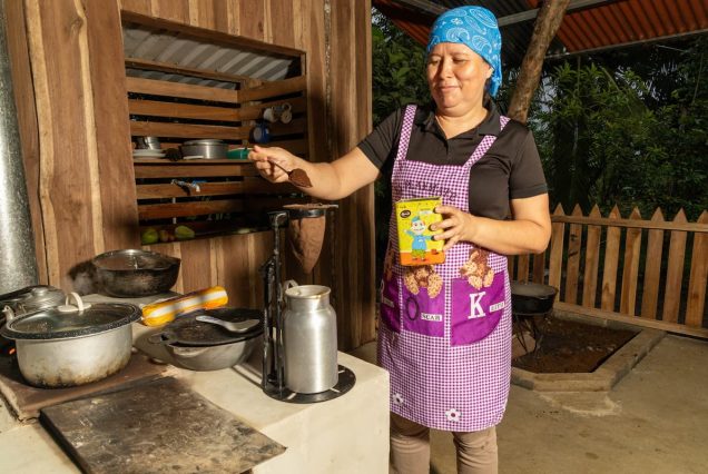 A woman in a purple apron pours chocolate powder into a cup in an outdoor kitchen with wooden walls.