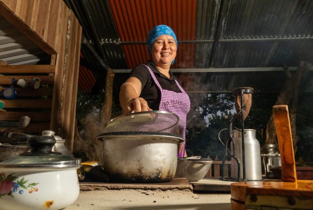 Person wearing a blue bandana and purple apron lifts the lid of a steaming pot in a rustic kitchen setting.