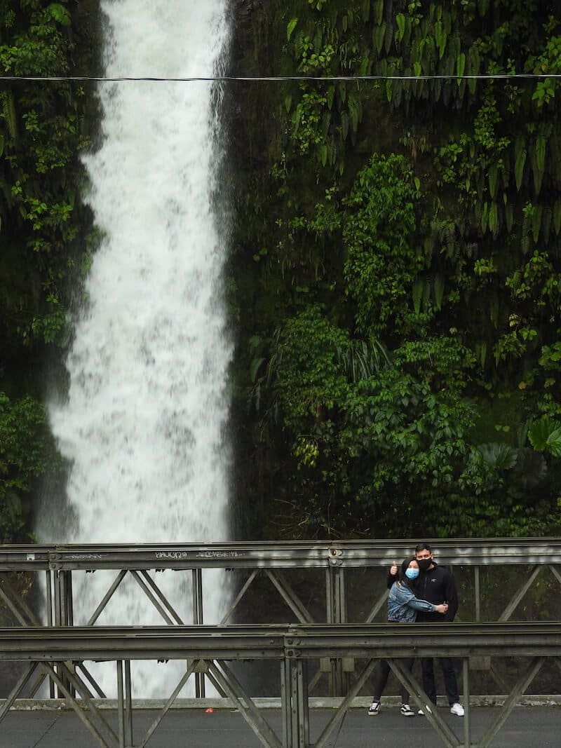 A couple posing for a photo on a bridge with a waterfall behind of them