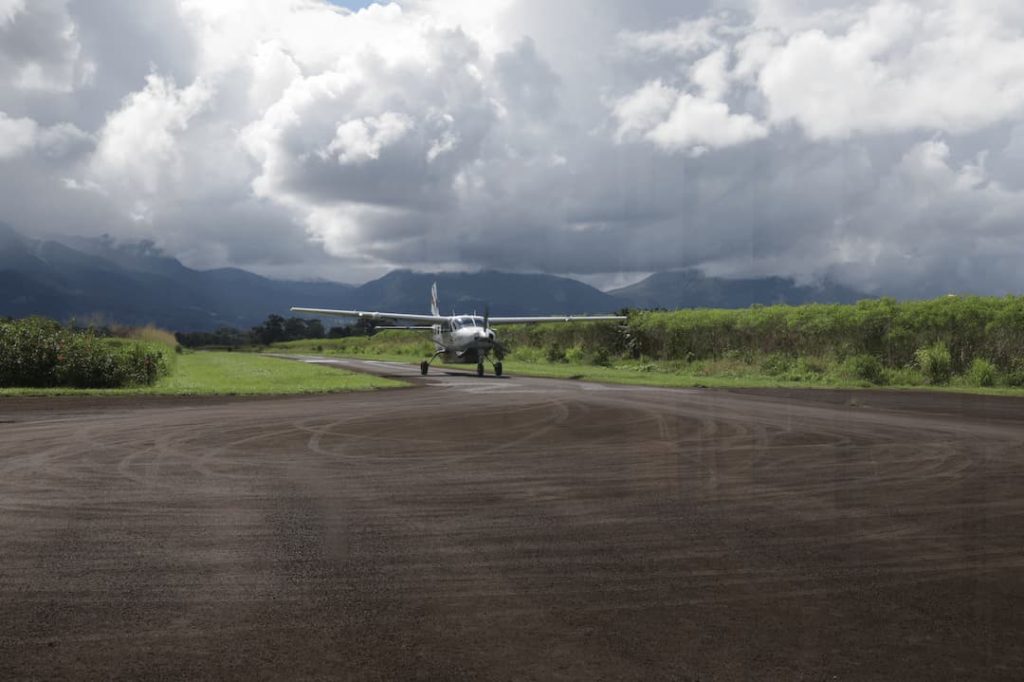 A small plane is stationed on a tarmac at La Fortuna Airport, surrounded by lush greenery under a cloudy sky, with mountains in the background.