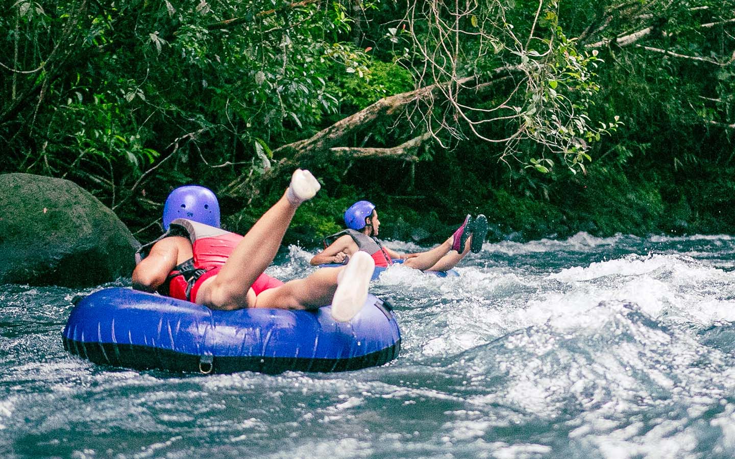 Two individuals are river tubing, battling through the choppy waters of a lush river. The forefront individual, wearing a red life vest and a blue helmet, has their arms raised high, seemingly enjoying the thrill. Behind them, a second individual in a similar protective gear appears focused on navigating through the rough river. Surrounding them, the river is lined with dense greenery, enhancing the sense of adventure in the wild