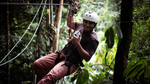 A person wearing a white helmet and brown clothing is ziplining through a lush foresting a Costa Rica adventures. They are attached to a cable with a harness and are holding onto a device that controls their movement. The greenery of the forest surrounds them, with tall trees and vibrant leaves in the background.