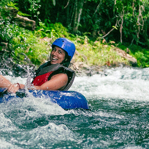 A person wearing a helmet and life vest beams with joy while tubing down the vibrant waters of Rio Celeste, surrounded by lush greenery.