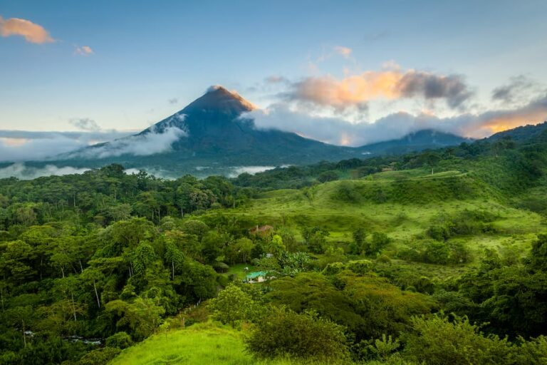 A lush green forest landscape in Costa Rica, with a prominent, cloud-covered, cone-shaped volcano in the background under a sky with scattered clouds at sunrise or sunset—truly a stunning scene typical of October weather.
