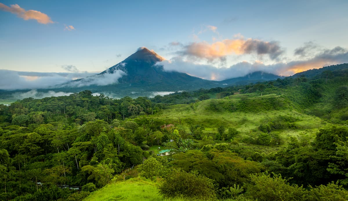 A lush green forest landscape in Costa Rica, with a prominent, cloud-covered, cone-shaped volcano in the background under a sky with scattered clouds at sunrise or sunset—truly a stunning scene typical of October weather.