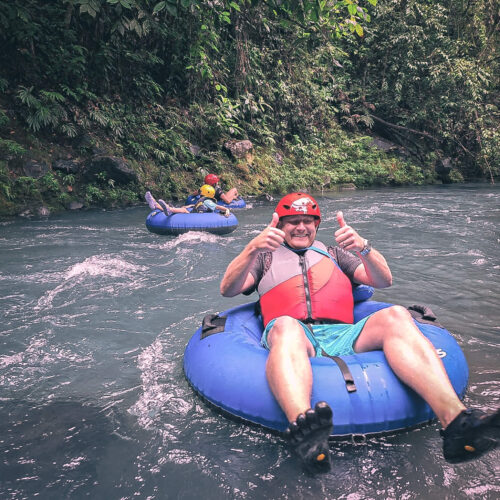 A man gives a thumbs-up while enjoying a tubing excursion on a river, wearing a red helmet and a life vest. In the background, another participant is lounging on a blue tube, both surrounded by the lush foliage of a tropical forest.