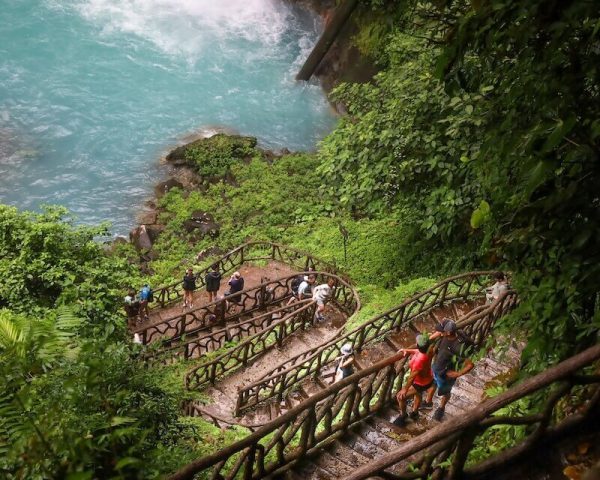 A group of people are walking up stairs to a waterfall in the Rio Celeste location.
