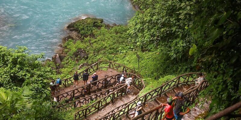 A group of people are walking up stairs to a waterfall in the Rio Celeste location.
