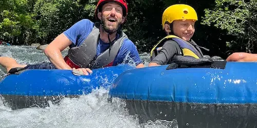 A man and a child, wearing helmets and life vests, are tubing down the mesmerizing waters of Rio Celeste, enjoying the lush greenery around them. A man and a child, wearing helmets and life vests, are tubing down the mesmerizing waters of Rio Celeste, enjoying the lush greenery around them.