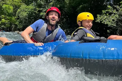 A man and a child, wearing helmets and life vests, are tubing down the mesmerizing waters of Rio Celeste, enjoying the lush greenery around them. A man and a child, wearing helmets and life vests, are tubing down the mesmerizing waters of Rio Celeste, enjoying the lush greenery around them.
