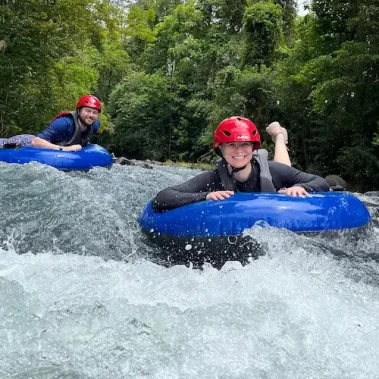 Two people wearing helmets smile as they float down the Rio Celeste on blue inner tubes, surrounded by lush green trees.