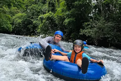 Two people wearing helmets and life jackets are tubing down Rio Celeste, surrounded by lush green trees, smiling as they enjoy the ride.