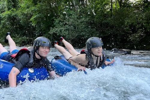 Two people wearing helmets and life jackets ride blue inner tubes down a river in a tropical paradise, surrounded by lush greenery, enjoying the warm August weather. Two people wearing helmets and life jackets ride blue inner tubes down a river in a tropical paradise, surrounded by lush greenery, enjoying the warm August weather.