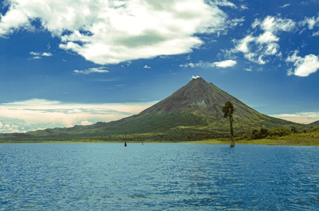 A distant, conical volcano with a smoke plume stands against a blue sky with clouds. In the foreground, a calm blue body of water meets a lush green shoreline, reminiscent of Costa Rica travel with its pristine beaches and lush rainforests.