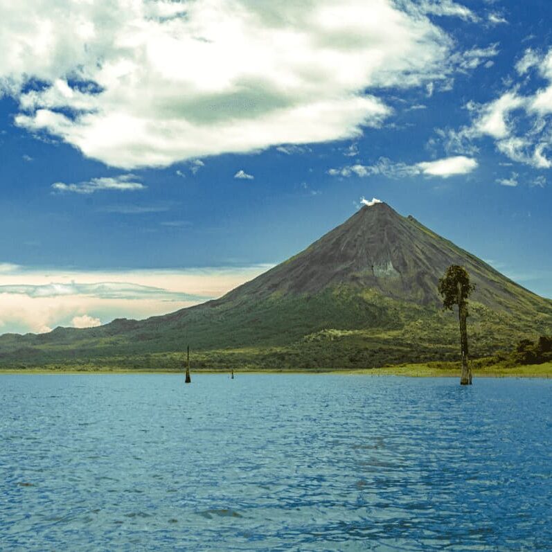 A distant, conical volcano with a smoke plume stands against a blue sky with clouds. In the foreground, a calm blue body of water meets a lush green shoreline, reminiscent of Costa Rica travel with its pristine beaches and lush rainforests.