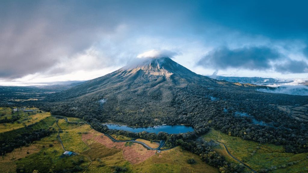 Aerial view of Volcan Arenal, a large, forested volcano with a cloud-covered peak, surrounded by lush green terrain, a winding road, and a small lake in the foreground.