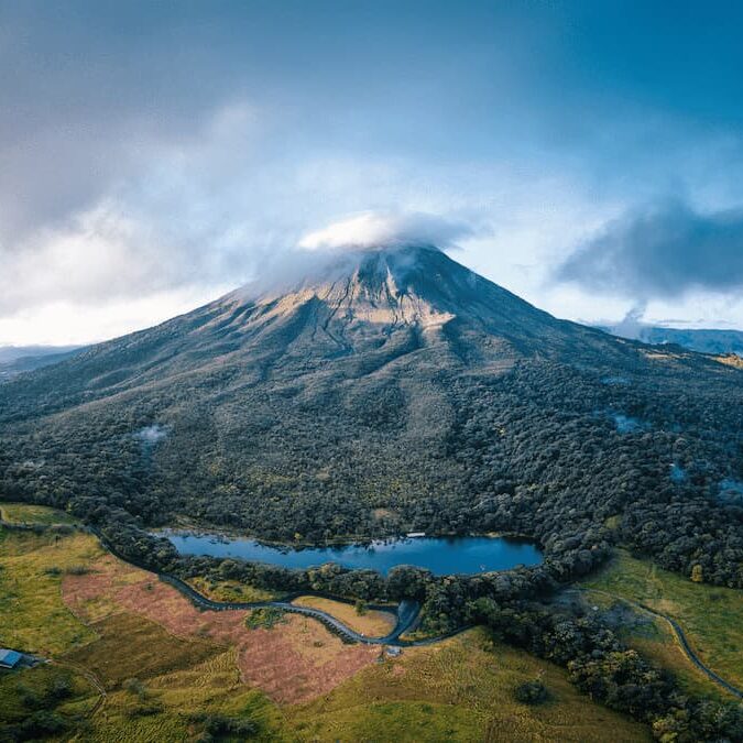 Aerial view of Arenal Volcano, a large volcanic mountain partially covered by clouds, surrounded by dense forest, a lake, and green open fields.
