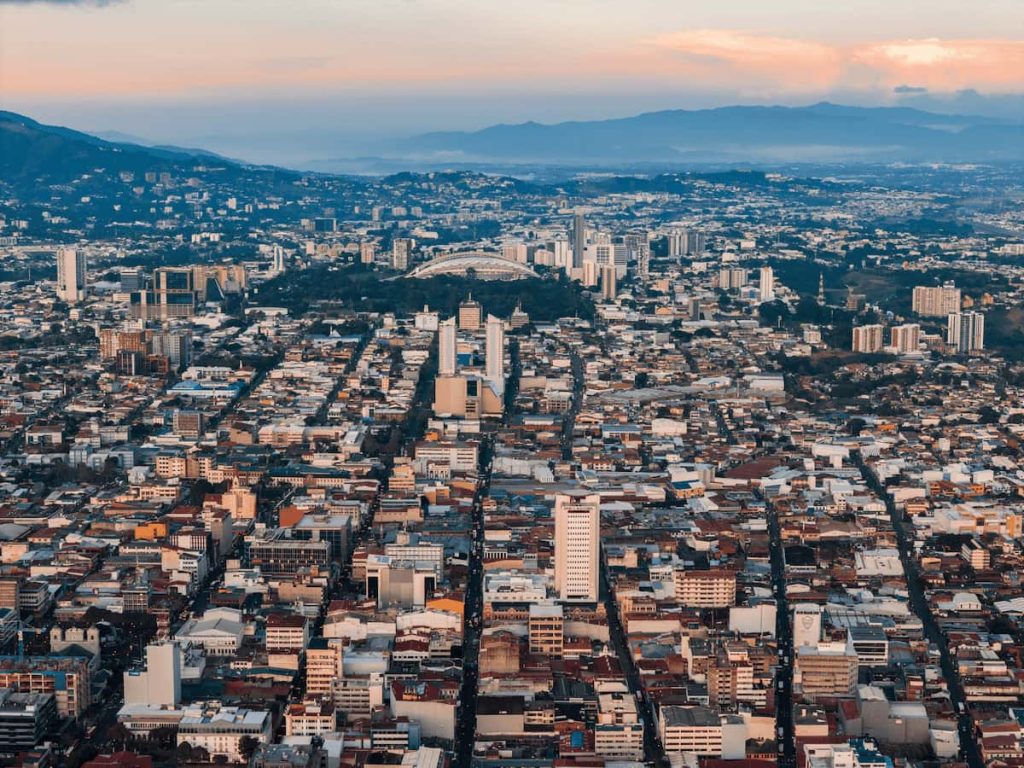 Aerial view of a densely populated city in Costa Rica with numerous buildings, roads, and surrounding mountains under a partly cloudy sky, offering a blend of urban life and lush rainforests.