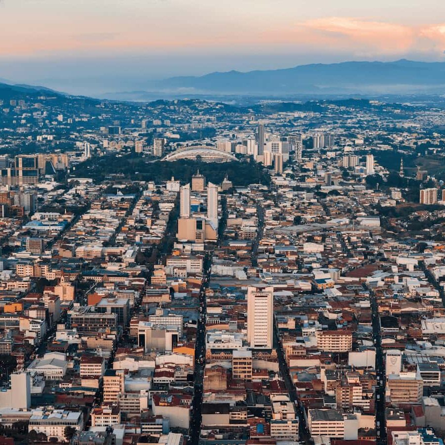 Aerial view of a densely populated city in Costa Rica with numerous buildings, roads, and surrounding mountains under a partly cloudy sky, offering a blend of urban life and lush rainforests.