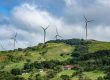 Wind turbines harness renewable energy on a lush green hillside in Costa Rica, with scattered houses and a cloudy sky as the backdrop.