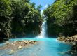 The Rio Celeste Waterfall flows into a vibrant blue river surrounded by lush green foliage under a clear blue sky.