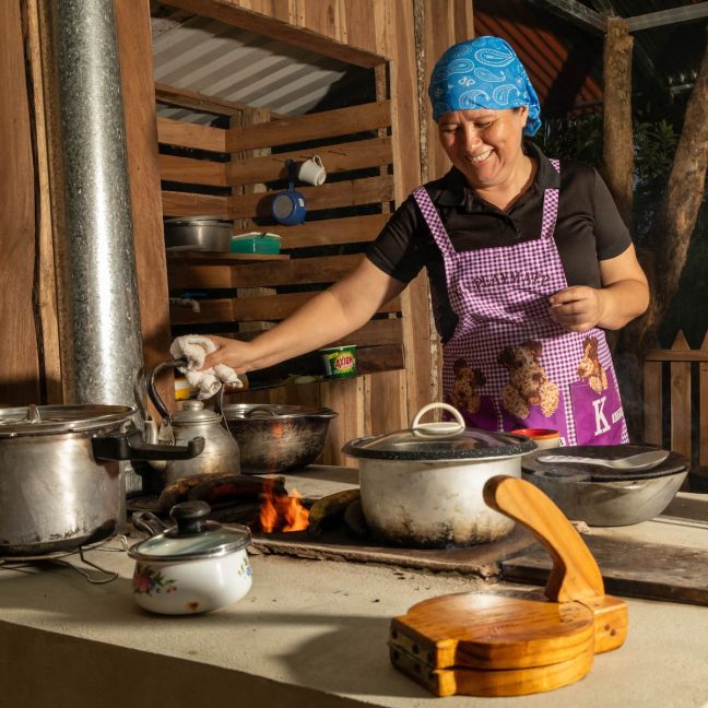 A woman in an apron and blue bandana is cooking at an outdoor stove with multiple pots and pans. Smiling as she handles a kettle, she's preparing a feast after an exhilarating day at Adventure Park Costa Rica, where zip lining through the jungle was the highlight.