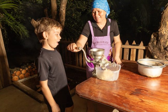 A woman in a purple apron uses a manual grinder at a wooden table, while a boy watches with excitement, reminiscent of the thrilling anticipation before zip lining in Costa Rica. A container collects the ground food as trees stand tall in the background like those surrounding an adventure park.