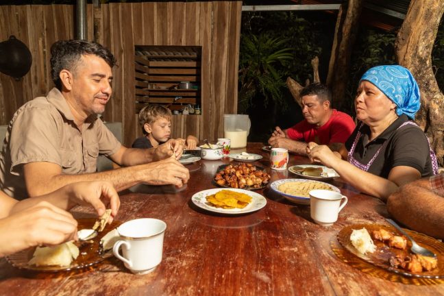 A family sits around a wooden table enjoying a meal with various dishes, including plantains and sauce, outside in a rustic setting, reminiscent of a Costa Rica cooking class.