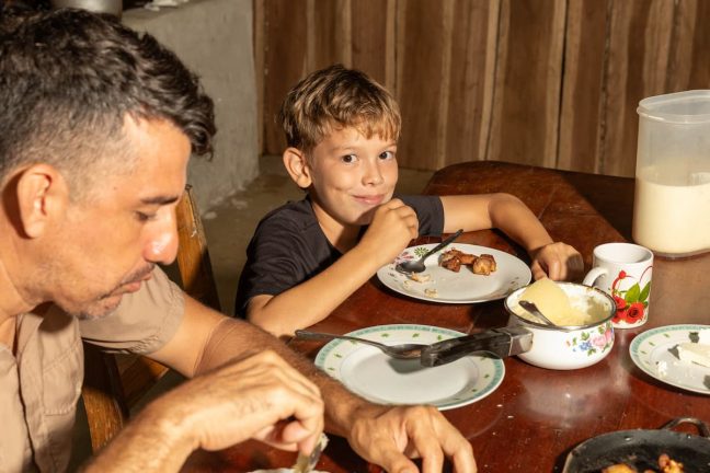A man and a boy are seated at a wooden table, eating a meal after an exciting day zip lining in Costa Rica. The boy, smiling, holds a fork with food on his plate. Dishes and a jug are on the table as they recall their thrilling adventure.
