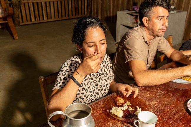 A woman in a leopard print shirt enjoys her meal from a plate at a wooden table near Rio Celeste, while a man sits beside her. A silver pitcher and cups are also on the table, reflecting the thrill of their day zip-lining through Costa Rica's lush adventure parks.