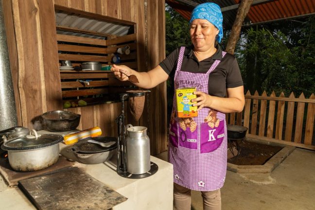 A woman in an apron prepares food in an outdoor kitchen at Adventure Park Costa Rica. Holding a small box, she stirs contents in a pot on the stove, surrounded by cooking utensils—fueling up for a day of excitement on the zip lining and ropes course nearby.