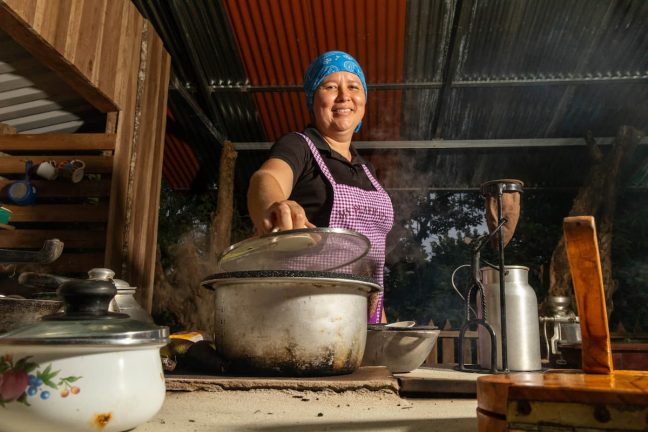 In an outdoor kitchen, a woman in a blue headscarf and purple apron lifts the lid from a steaming pot, reminiscent of the adventurous spirit at an Adventure Park in Costa Rica.