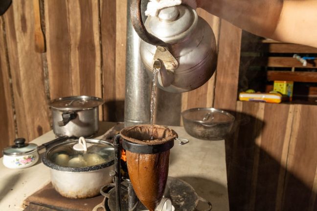 Hand pouring hot water from a kettle into a cloth filter set on a wooden stand, near pots and a stove in this rustic kitchen setting evokes the same thrill as experiencing the challenge of traversing Rio Celeste's rugged paths.