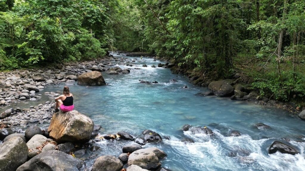 A person sits on a large rock beside the electric blue color of Rio Celeste, surrounded by dense green forest and scattered rocks.