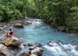 A person sits on a large rock beside the electric blue color of Rio Celeste, surrounded by dense green forest and scattered rocks.