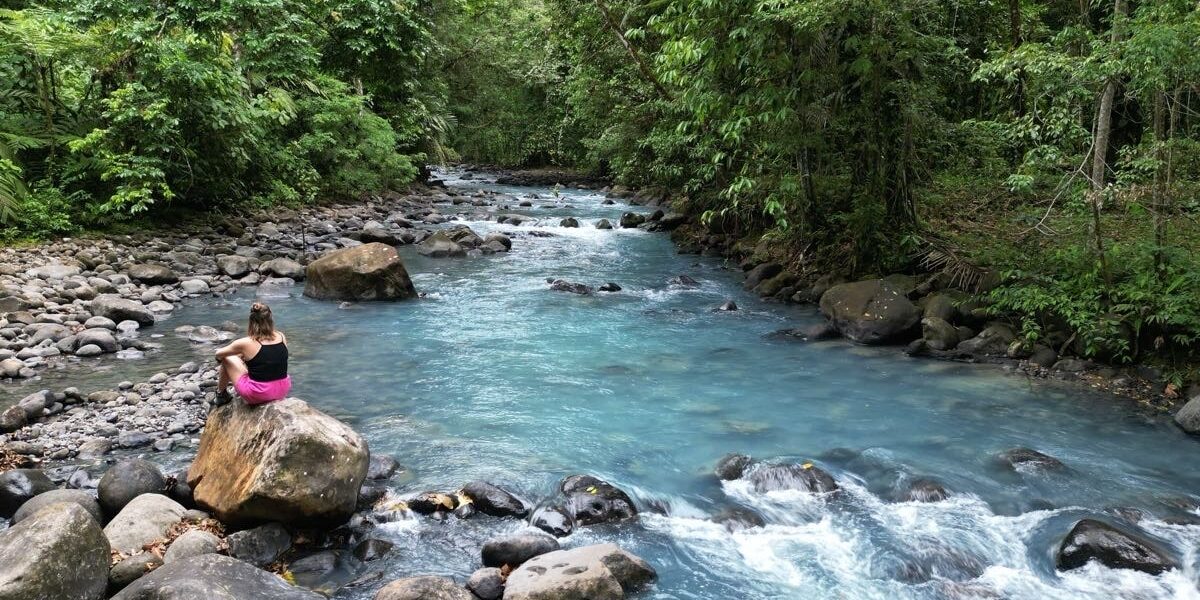A person sits on a large rock beside the electric blue color of Rio Celeste, surrounded by dense green forest and scattered rocks.