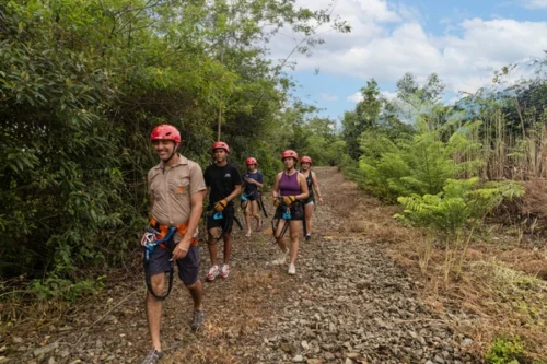 A group of people wearing helmets and harnesses walk on a rocky path surrounded by greenery and trees, heading outdoors with Onca Tours, showcasing their certifications in adventure safety.